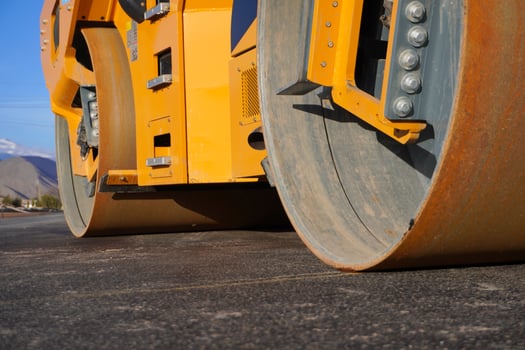 A close-up view of a yellow road roller, showcasing its large, steel wheels with a rusty finish. The background features a clear blue sky and distant mountains. photo