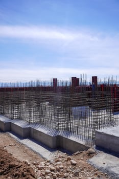 A construction site featuring a grid of steel rebar protruding from concrete foundations. The background shows red formwork against a bright blue sky. photo