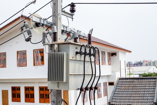 A distribution transformer mounted on a power line pole is located among residential buildings, providing electricity to the entire area photo