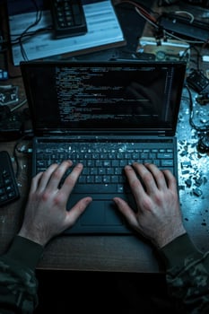 Young man typing code on laptop surrounded by tech gadgets at a cluttered desk photo