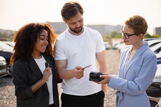 A diverse couple purchasing a new car, paying with a credit card, and receiving keys from a friendly saleswoman at a dealership. photo