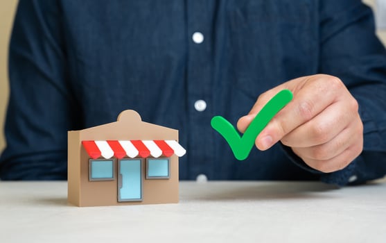 A man holds a green check mark near a store. Pass the test. Quality verification. Business permits. Successful inspection, business licensing, or compliance with regulations. Audit approval photo