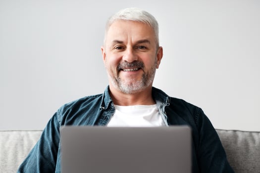 Cheerful middle-aged grey-haired man using laptop while resting on sofa in living room, empty space. Handsome mature man reading online blog or chatting, surfing on Internet at home, closeup photo