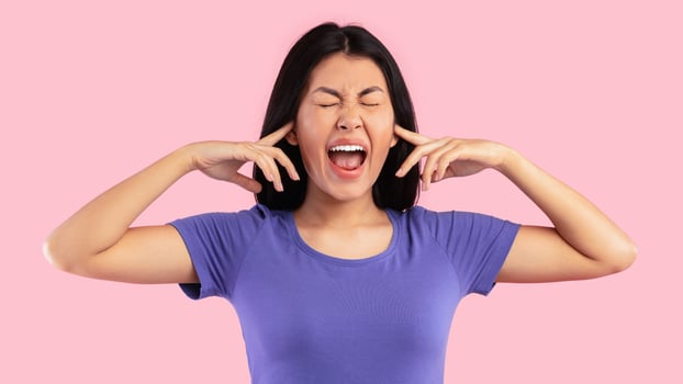 I Don't Want To Hear It. Portrait of irritated young asian woman covering her ears, plugging with fingers and screaming in anger. Upset lady standing isolated over pink studio background. Noisy sound photo