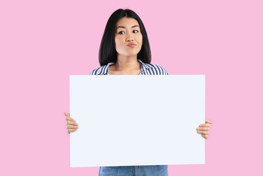Making Decision. Portrait of doubtful asian woman holding empty blank board isolated on pink studio background. Unsure lady standing with white square paper placard for ad, thinking and looking away photo