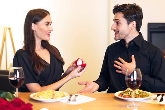 Will You Marry Me. Smiling millennial woman proposing to her boyfriend, holding and showing open box, offering marriage ring to surprised man, getting engaged in restaurant during romantic date photo
