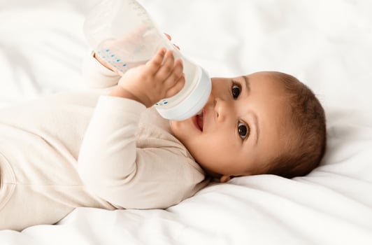 Childcare And Nutrition Concept. Portrait of cute little African American baby drinking from bottle, wearing bodysuit lying on the white blanket at home. Black infant child on bed in the bedroom photo