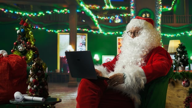Santa Claus reads letters from kids on laptop and replying in Christmas ornaments adorn workshop. Old costumed man in holiday season ambiance reading children wish lists, typing on notebook, camera A photo