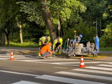 Four workers apply fresh paint to crosswalk lines on a quiet road bordered by trees in a city park, showing teamwork and effort photo