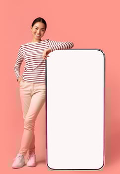 Mobile Offer. Beautiful Smiling Asian Woman Leaning At Big Smartphone With Blank White Screen, Showing Free Copy Space For Your Design Or Advertisement, Posing Over Pink Background, Mockup Image photo