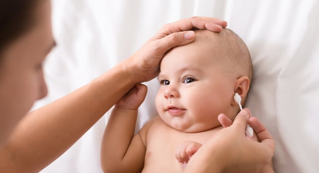 Newborn hygiene. Mother cleaning baby ear with cotton swab, infant lying and looking at her, copy space photo