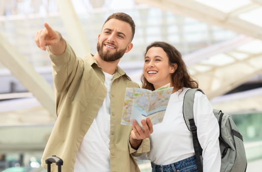 Travelling Concept. Happy Young Couple With City Map Standing Near Airport Building After Arrival To New Country, Cheerful Millennial Man And Woman Planning Travel Route, Man Pointing Away photo
