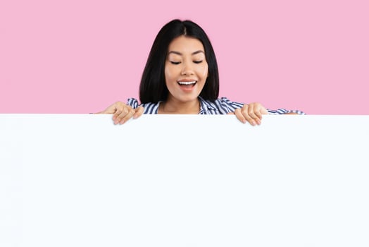 Place For Your Promo. Excited asian woman standing behind white empty board for advertisement over pink background. Lady peeking out big blank advertisement panel, looking down at empty space for text photo