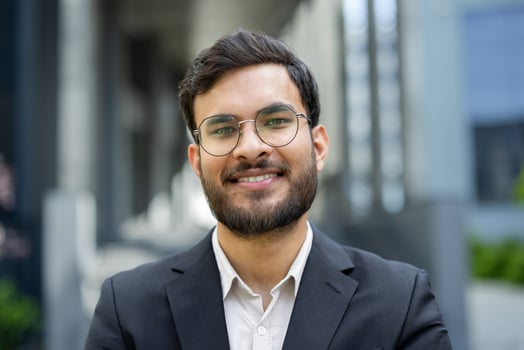 A smiling businessman with a beard and glasses stands confidently outdoors in front of a building, wearing a suit. photo