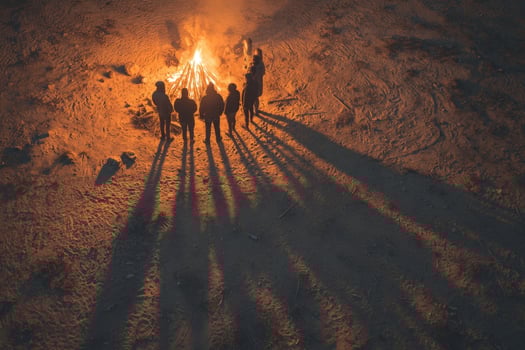 Group of friends gathered around a campfire at dusk, casting long shadows on sandy ground photo