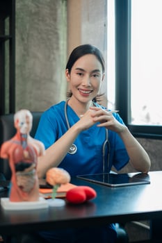 Smiling female doctor in blue scrubs at desk with medical models and tablet, conveying professionalism and healthcare expertise. photo