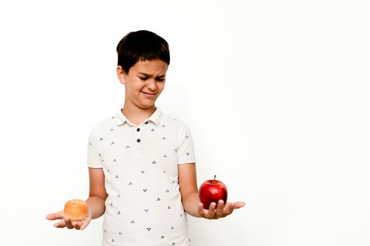 A young boy in a white shirt stands isolated on a white background, holding an apple in one hand and a donut in the other, with a conflicted expression. Suitable for illustrating healthy eating choices, nutrition education, and food decision concepts. photo