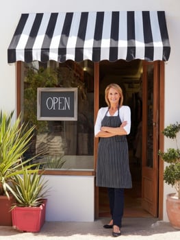Female shop owner standing outside store entrance with open sign and striped awning, smiling confidently in apron photo