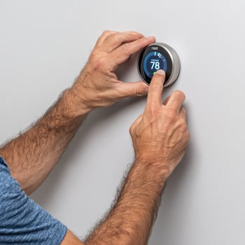 Smart thermostat on wall being adjusted by man with hairy arms, showing temperature 78 degrees in blue digital display photo