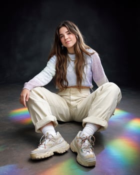 Young woman with long hair sitting on floor wearing pastel sweater and beige pants with colorful light reflections creating joyful mood photo