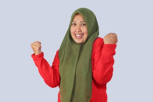 Joyful hijab woman making victory gesture with both fists, expressing excitement, motivation, and achievement, isolated studio photo for positive success themes