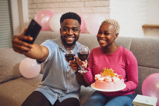 Young African couple sharing a birthday cake with lit candles, capturing the special moment on a smartphone selfie. photo