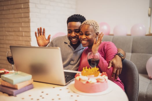 An affectionate couple shares a virtual birthday celebration with loved ones, enjoying cake and wine while connecting over a computer in a cozy indoor setting photo