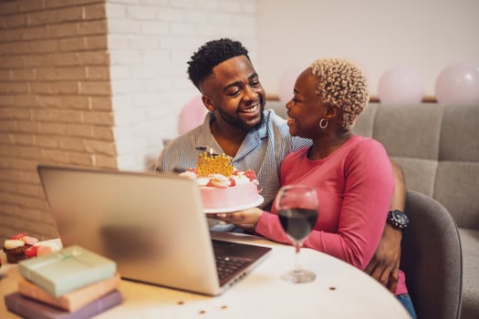 A young African American couple celebrating a birthday via call, smiling happily with a birthday cake and wine, sharing joy and love indoors. photo
