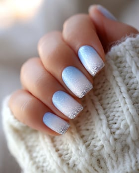 Hyperreal macro of elegant ombre nail gradient in cool winter tones icy blue fading to snow white glitter tips warm indoor lighting soft cozy texture of knit background feminine aesthetic photo