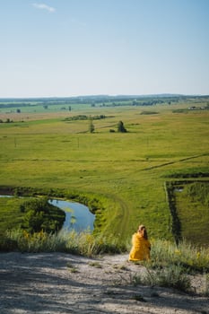A woman, dressed elegantly in a vibrant yellow dress, sits gracefully on a picturesque hill while enjoying the serene and beautiful view of the winding river on a lovely sunny day photo