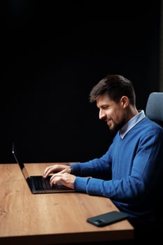 Man working on laptop at wooden desk during evening hours with focused expression photo