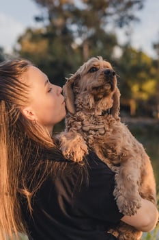 A young Caucasian woman with long brown hair holds a light brown cocker spaniel dog close to her face, sharing a moment of affection outdoors. photo