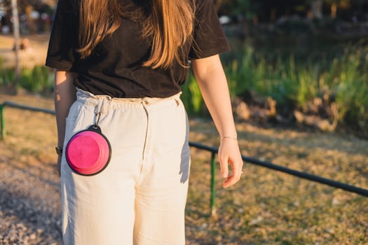 A young woman with long brown hair wears a black t-shirt and light-colored pants. A pink dog treat pouch hangs from her waist in a park setting. photo