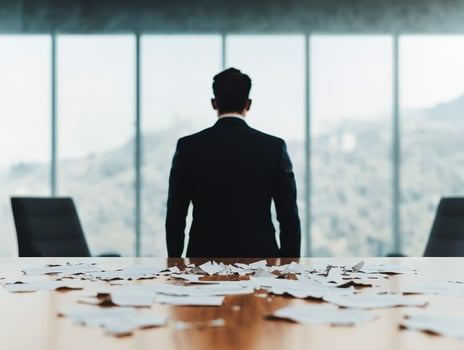 Businessman in suit stands facing large windows behind table covered with torn papers, expressing frustration and tension photo