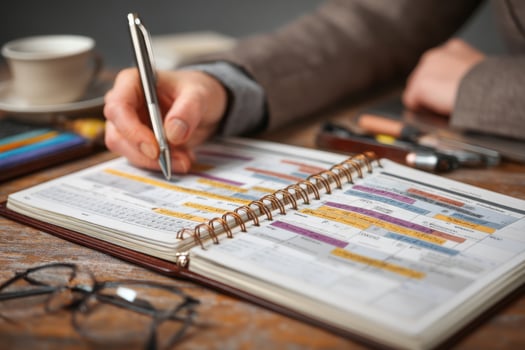A person writing in a planner on a desk with a cup of coffee and glasses photo