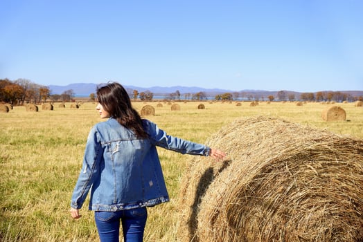 Young brunette woman in denim jacket standing near haystack in field with bales of hay. she looking to the left. Primorsky kray, Russia. Beauty in nature. Copy space photo