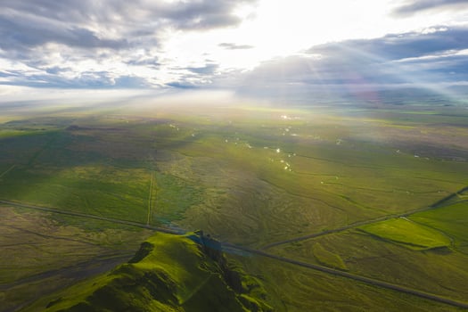 Aerial view of a green Icelandic landscape at sunset, featuring a grassy hill, scattered clouds, sunlight beams, a straight road, and small water bodies. photo