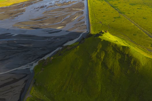 Aerial image of a green hill in golden sunlight, bordered by a dark braided river system and flat green fields with visible tracks in Iceland. photo