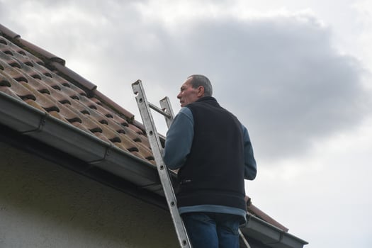 Senior roofer climbs an aluminum ladder, carefully inspecting the roof of a residential building under a cloudy sky, taking precautions during maintenance, photo