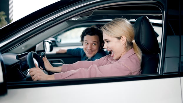 Young woman excitedly test drives a new car in a showroom, while a smiling man observes, showcasing the thrill of car shopping experience photo