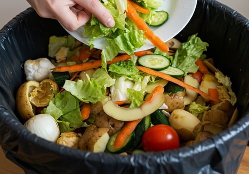 Person throwing salad in garbage can, kitchen background, illustrating food waste photo
