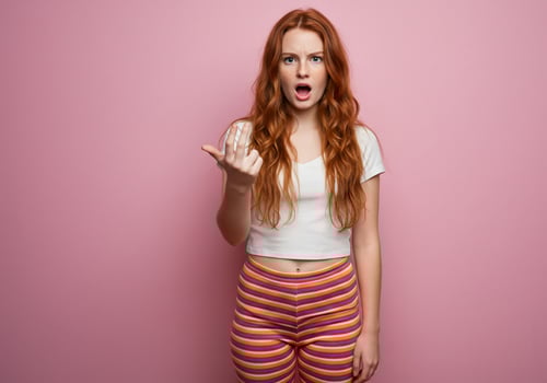 Redhead woman gestures with her hand against a pink background, looking shocked photo