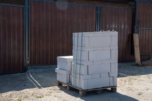 Stack of lightweight aerated concrete blocks secured with straps on wooden pallet at construction site ready to start construction, with wood grain metal fence in the background on a sunny day, photo