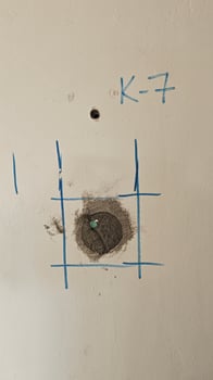 A close-up photo showing a concrete core sample taken from a building wall for an earthquake resistance test. The K-7 mark on the wall is a notable detail.