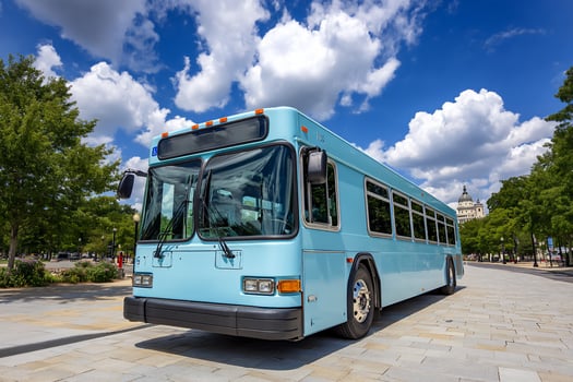A light blue bus on a paved road near trees under a blue sky with white clouds photo