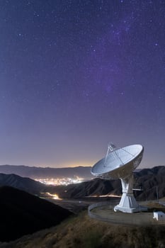 Antenna sits on a hill overlooking a city under a starry, dark-blue night sky photo