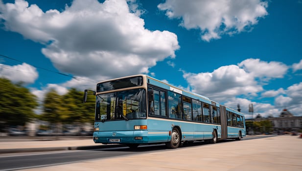 Blue articulated bus is driving on a street on a sunny day with clouds and trees photo