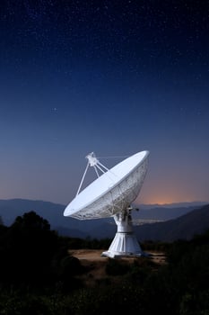Satellite dish against a starry sky. Trees and hills dot the landscape photo