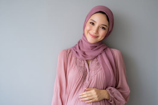 Pregnant smiling woman with hijab, holding belly against grey wall in studio photo