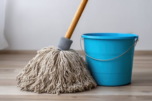 Mop with a wood handle and blue bucket sits on a light wood floor by a white wall photo